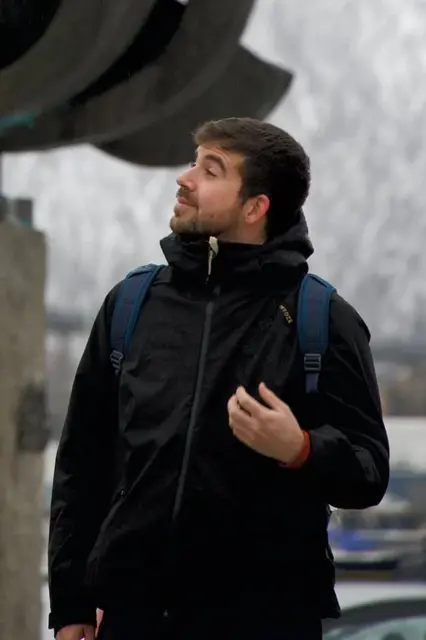 Manuel looking to the side with a backpack in Tromsø, Norway. He is wearing a black jacket and a blue backpack.